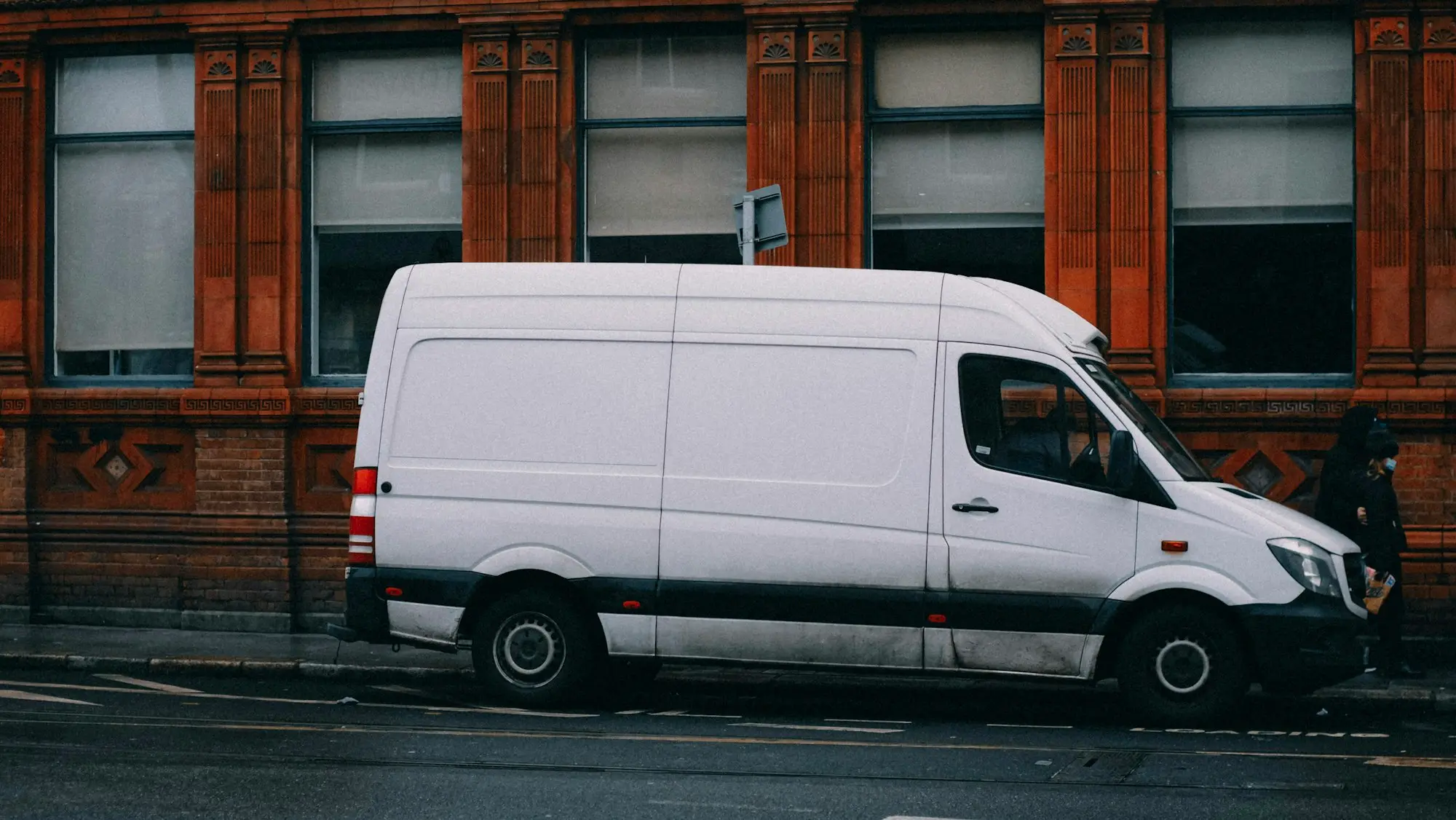 White high-roof panel van parked at the kerb beside a brick building