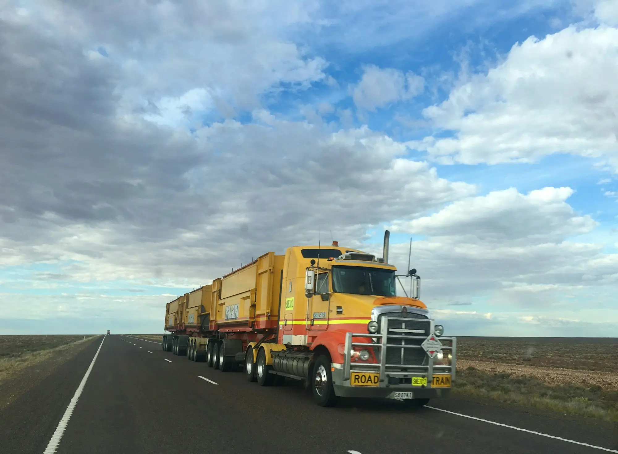 Road train hauling multiple trailers along a straight outback highway under a wide cloudy sky