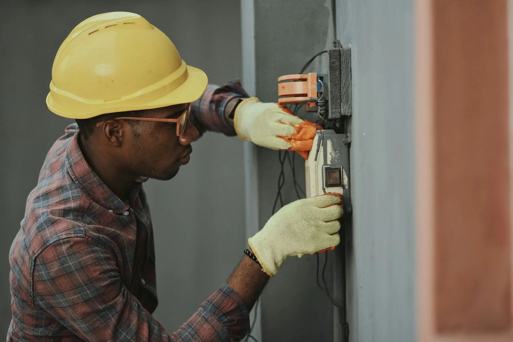Electrician in a yellow hard hat and gloves installing wiring at a meter box